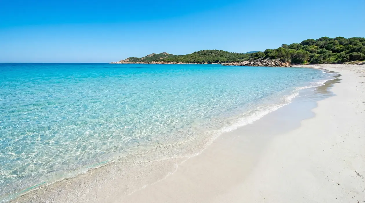 Spiaggia di sabbia bianca con mare turchese e acqua trasparente, costa verde sullo sfondo in una giornata serena
