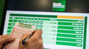 Mano che compila una schedina del SuperEnalotto davanti a uno schermo con risultati e quote della vincita