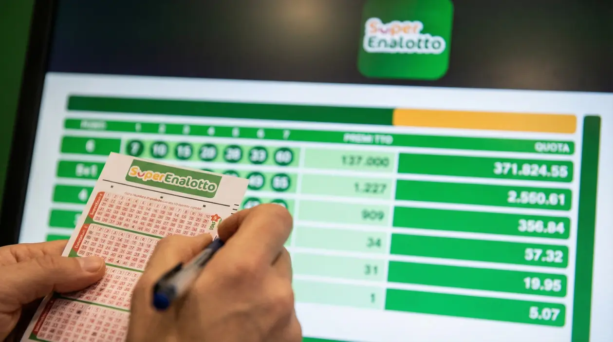 Mano che compila una schedina del SuperEnalotto davanti a uno schermo con risultati e quote della vincita